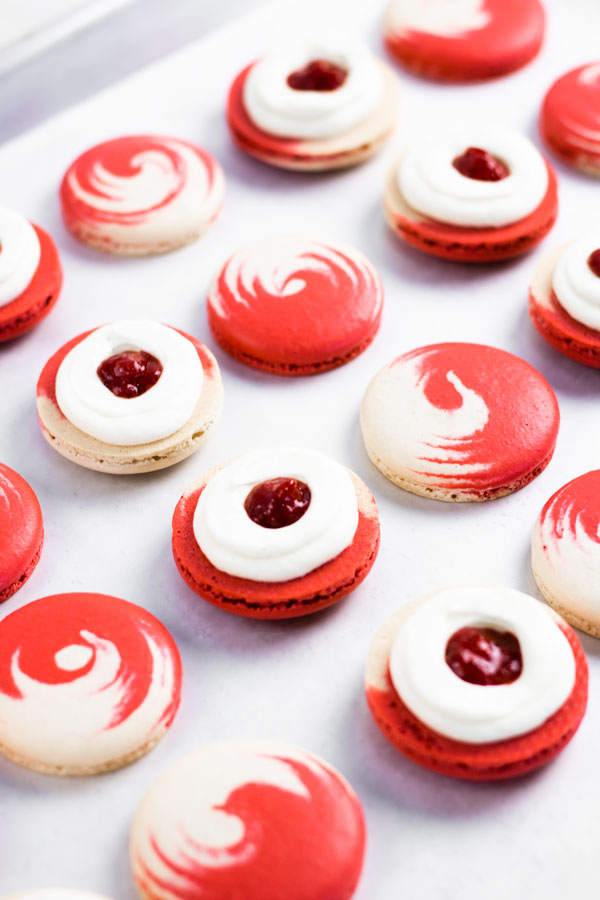 red and white macaron shells with cream cheese and strawberry jam filling on baking sheet