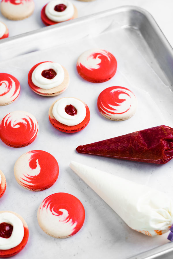 red and white macaron shells with pastry bags filled with cream cheese and strawberry jam filling on baking sheet