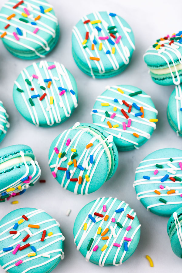 turquoise macarons with cream cheese filling with white drizzle and colorful sprinkles scattered on white background