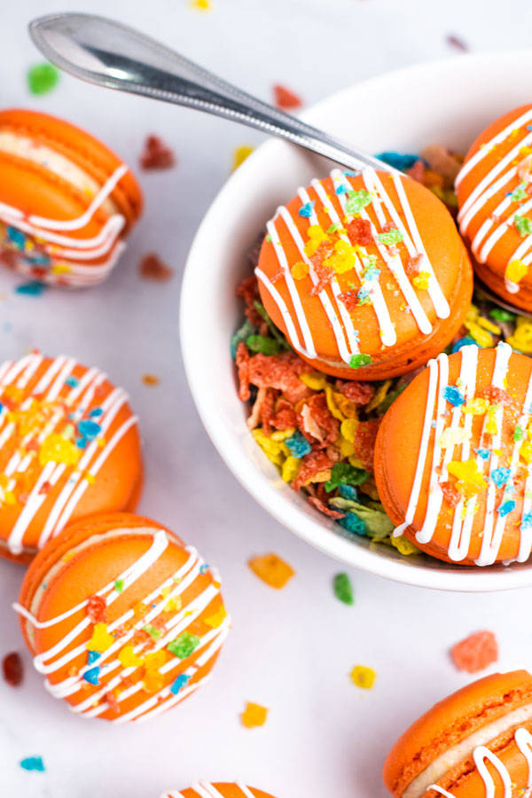 orange fruity pebbles macarons with white drizzle and cereal sprinkles inside cereal bowl