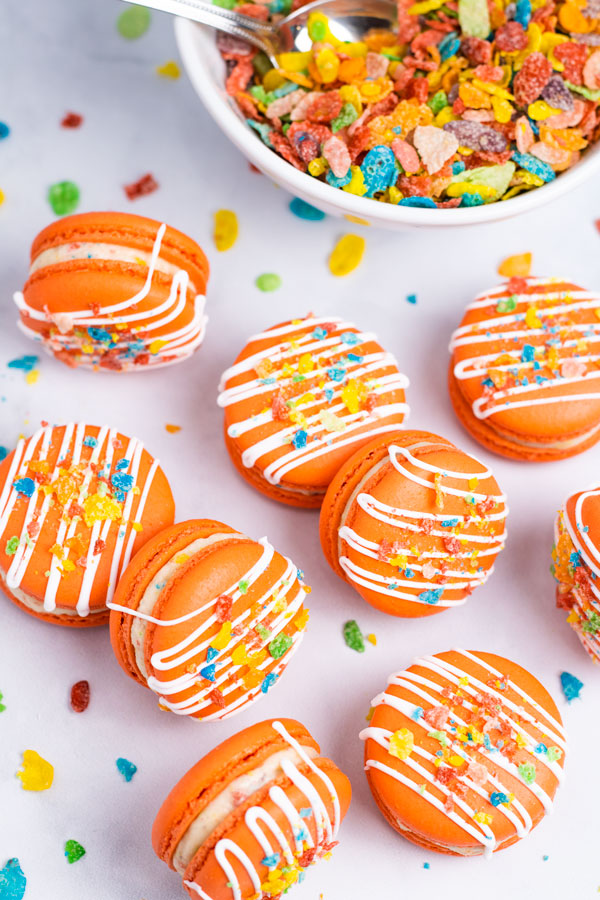 orange fruity pebbles macarons with white drizzle and cereal sprinkles next to cereal bowl