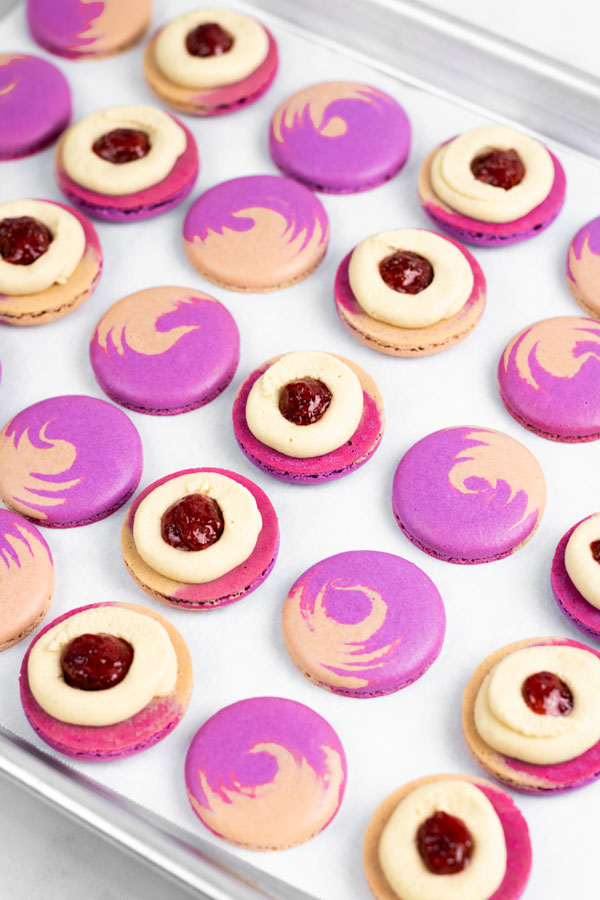 purple and brown peanut butter and jelly macaron shells on baking sheet with buttercream