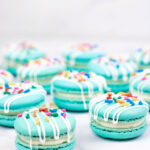 turquoise macarons with cream cheese filling with white drizzle and colorful sprinkles on white background
