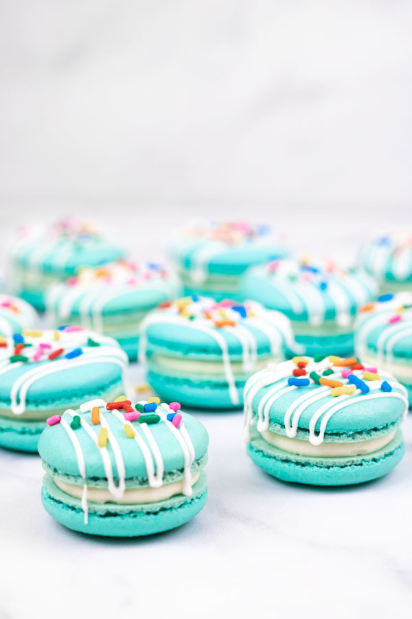 turquoise macarons with cream cheese filling with white drizzle and colorful sprinkles on white background