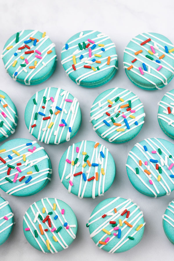 turquoise macarons with cream cheese filling with white drizzle and colorful sprinkles on white background