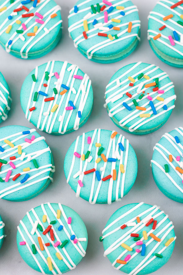 closeup of turquoise macarons with cream cheese filling with white drizzle and colorful sprinkles on white background