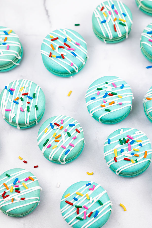 turquoise macarons with cream cheese filling with white drizzle and colorful sprinkles on white background