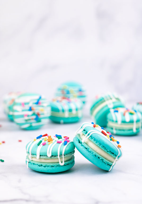 Two turquoise macarons with cream cheese filling with white drizzle and colorful sprinkles in front of macarons and white background