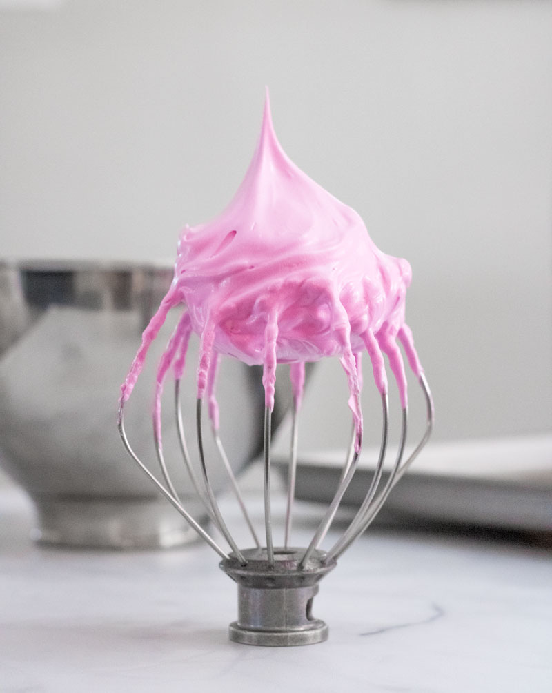 whisk with pink meringue in front of stainless steel mixingbowl