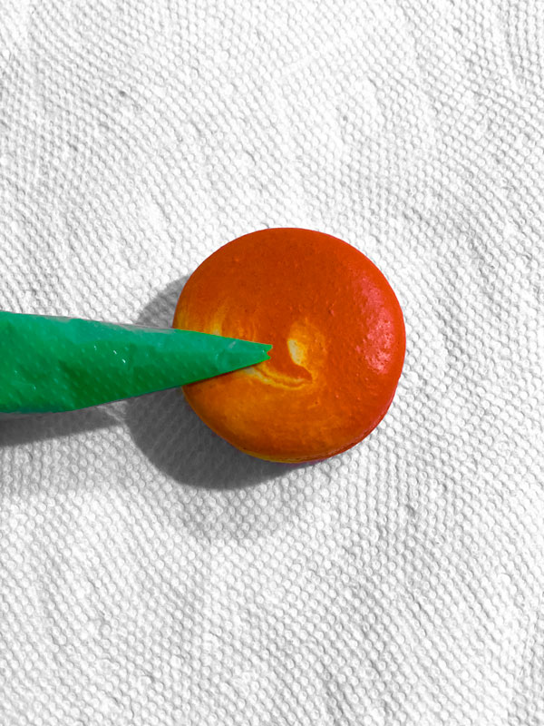 piping bag with green batter on top of yellow and orange swirled peach macaron