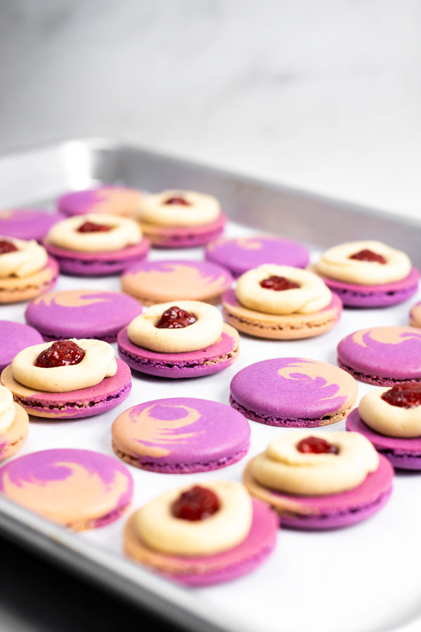 purple and brown peanut butter and jelly macaron shells on baking sheet with buttercream and jelly